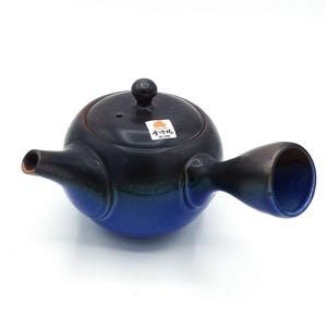 Può includere: Una teiera in ceramica blu e nera con un corpo arrotondato e un beccuccio laterale. La teiera ha un piccolo coperchio con un pomo in cima e un manico che si estende lateralmente. La teiera ha un'etichetta sul lato che dice "小粋茶" in giapponese.
