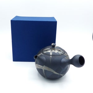 Op de afbeelding: Een zwarte theepot van keramiek met een zilveren glazuurontwerp. De theepot heeft een rond lichaam en een korte tuit. De theepot staat op een blauwe doos.