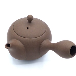 May include: A brown ceramic teapot with a rounded body and a curved spout. The teapot has a small knob on the lid.