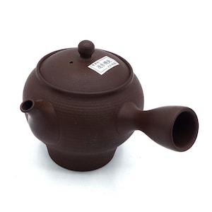 Può includere: Una teiera in ceramica marrone con un beccuccio corto e un manico che si curva verso l'alto e lontano dal corpo. La teiera ha un'etichetta sul coperchio che recita "茶器  益寿窯  大西陶芸".