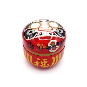 Könnte beinhalten: Roter und goldener japanischer Glücksbringer mit einem lächelnden Gesicht und dem Wort "福" (bedeutet "Glück") auf der Unterseite.