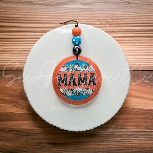 May include: A round orange and blue patterned ornament with the word "MAMA" in black lettering. The ornament is hanging from a string with orange, blue, and black beads.