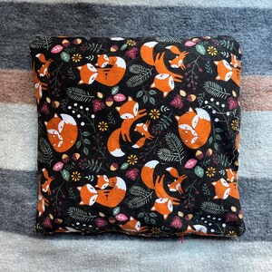 May include: A black and orange pillow with a repeating pattern of foxes and leaves. The foxes are orange with white accents and are holding a smaller fox.