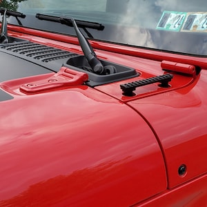 May include: Close-up of a red Jeep Wrangler's hood with black accents. Features include windshield wipers, a black air vent, and a black rail. The hood's glossy finish reflects the surroundings.