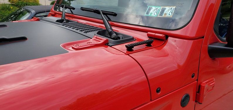 May include: Close-up of a red Jeep Wrangler's hood with black accents. Features include windshield wipers, a black air vent, and a black rail. The hood's glossy finish reflects the surroundings.