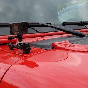 May include: A black action camera mounted on a black and silver bracket, attached to the red hood of a vehicle. The hood also features black windshield wipers and a red latch.