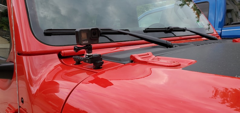 May include: A black action camera mounted on a black and silver bracket, attached to the red hood of a vehicle. The hood also features black windshield wipers and a red latch.