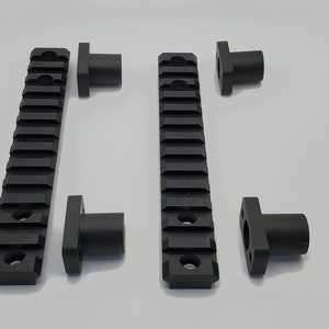 May include: Black metal Picatinny rails and mounting hardware. The rails have a series of horizontal slots for attaching accessories. The mounting hardware is cylindrical with a square base. The items are arranged on a white surface.