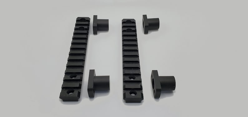 May include: Black metal Picatinny rails and mounting hardware. The rails have a series of horizontal slots for attaching accessories. The mounting hardware is cylindrical with a square base. The items are arranged on a white surface.
