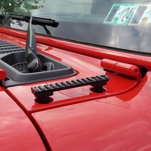 May include: Close-up of a red vehicle hood with a black accessory rail and red hinges. The hood features a black vent and windshield wipers. The paint is glossy, reflecting the surroundings. The image showcases automotive parts and accessories.