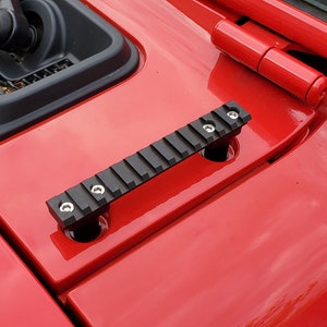 May include: Black metal accessory with a ridged top, mounted on a red vehicle hood. The accessory is secured with silver screws. The hood also features a red hinge and a black vent.
