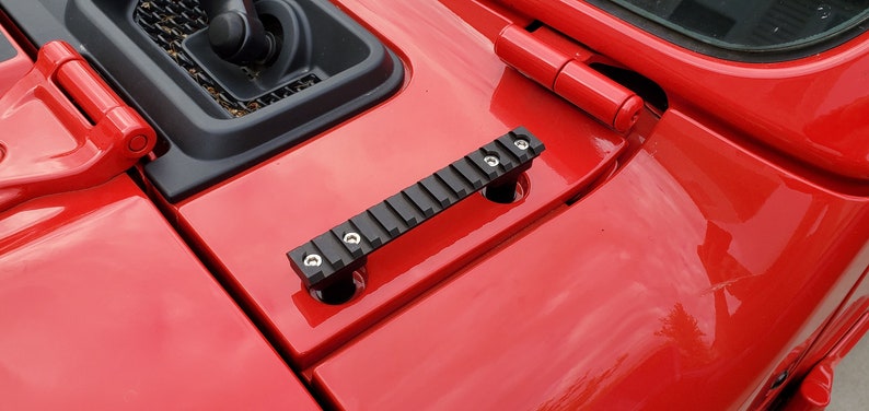 May include: Black metal accessory with a ridged top, mounted on a red vehicle hood. The accessory is secured with silver screws. The hood also features a red hinge and a black vent.