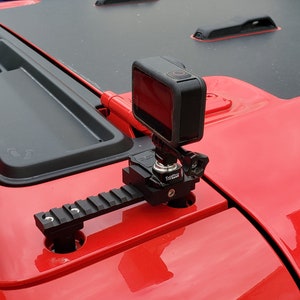 May include: A black action camera mounted on a black rail system, attached to the red hood of a vehicle. The camera is square-shaped and black. The rail system is mounted on the hood's surface.