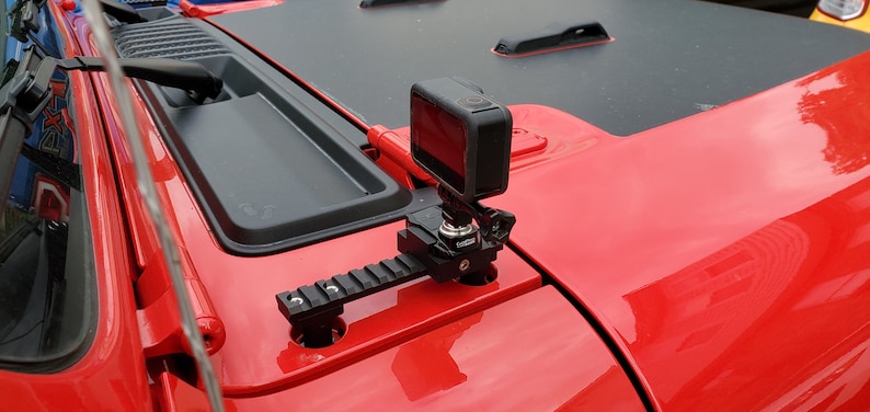 May include: A black action camera mounted on a black rail system, attached to the red hood of a vehicle. The camera is square-shaped and black. The rail system is mounted on the hood's surface.