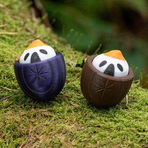 May include: Two decorative figurines, each featuring a white face with black eyes and a yellow-orange top, nestled in a purple and brown container. The containers are decorated with a starburst design. The figurines are set on a bed of green moss.