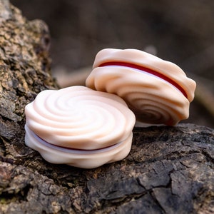 Puede incluir: Dos galletas de espiral rosa y blanca con relleno rojo, sentadas sobre una superficie de corteza de árbol marrón rugosa.