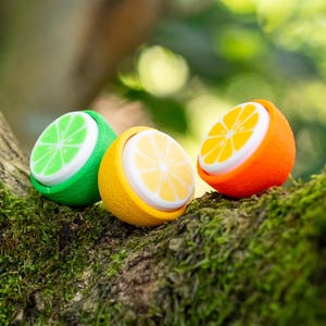 May include: Three small, round, fruit-shaped containers featuring textured peels in lime green, lemon yellow, and orange. Each container has a flat, white top printed with a stylised citrus slice graphic in a colour matching its respective outer shell.