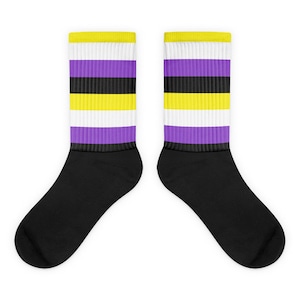 Peut inclure: Une paire de chaussettes noires avec des rayures jaunes, blanches, violettes et noires. Les rayures sont dans l'ordre du drapeau de la fierté non binaire.