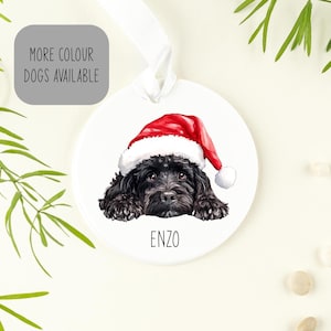 May include: A round white ceramic ornament featuring a black dog wearing a red and white Santa hat. The name ENZO is printed in black below the dog. A white ribbon is attached to the top for hanging. A grey box with the text MORE COLOUR DOGS AVAILABLE.