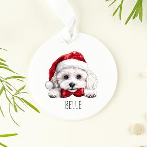 May include: White ceramic Christmas bauble featuring a white dog wearing a red Santa hat and a red bow tie. The dog's name, "Belle", is written beneath the dog.
