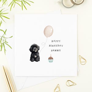 May include: A white birthday card featuring a black dog illustration, a beige balloon, and a cupcake. The text reads "HAPPY BIRTHDAY SAMMY". Gold paper clips and a gold pen are also visible, adding to the card's design.