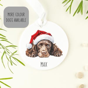 May include: A round white ceramic ornament featuring a brown spaniel dog wearing a red and white Santa hat. The name MAX is printed in black capital letters below the dog. A white ribbon is attached to the top for hanging the ornament.