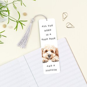 May include: White bookmark featuring a light brown and white dog illustration. The bookmark has the text "ALL YOU NEED IS A GOOD BOOK AND A POOCHON" and a silver tassel. It rests on a white notebook.