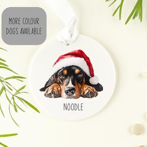 May include: White ceramic bauble featuring a black and brown dog wearing a red Santa hat. The dog is looking at the camera and the bauble says "NOODLE" at the bottom.