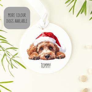 May include: A round white ceramic ornament featuring a watercolour illustration of a brown curly-haired dog wearing a red and white Santa hat. The name TOMMY is printed in black below the dog. A white ribbon is attached to the top for hanging.