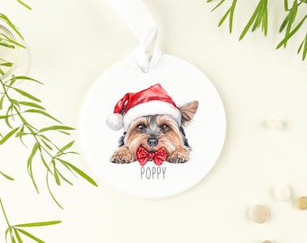 May include: White ceramic Christmas ornament featuring a Yorkshire Terrier wearing a Santa hat and a red bow tie. The dog's name, "Poppy", is written below the image.