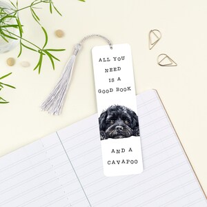 May include: A white bookmark featuring the text "ALL YOU NEED IS A GOOD BOOK AND A CAVAPOO" and a watercolour image of a black Cavapoo dog. The bookmark has a silver tassel and is on a notebook.