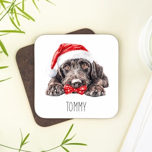 May include: A white ceramic coaster with a brown cork backing features a watercolour illustration of a brown dog wearing a red Santa hat and a red bow tie. The dog's name, "TOMMY", is printed in black below the illustration.