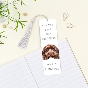 May include: White bookmark featuring a brown Cockapoo dog illustration and the text "ALL YOU NEED IS A GOOD BOOK AND A COCKAPOO". The bookmark has a silver tassel and is on a notebook.