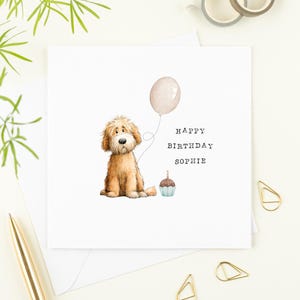 May include: A white birthday card with a cartoon dog, balloon, and cupcake illustration. The dog has a light brown, fluffy coat. The card reads "HAPPY BIRTHDAY SOPHIE".