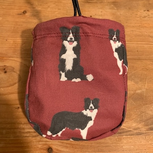May include: A burgundy drawstring bag with a black and white border collie dog print. The bag has a black drawstring and a black toggle.