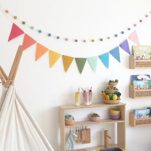 May include: A children's playroom with a white teepee, a wooden play kitchen, and rattan bookshelves. Colorful pennant banners and felt ball garlands decorate the white walls. The kitchen has a sink, stove, and storage. Books and toys are arranged throughout the room.