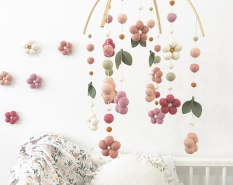 May include: A hanging mobile featuring felted wool spheres and flower shapes in shades of pink, white, and sage green, suspended from a curved wooden frame. Several matching felt flowers are attached to the wall behind the mobile.