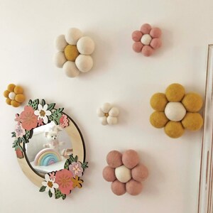 May include: A collection of felt flower wall decor in various sizes and colors, including white, yellow, and pink. A wooden framed mirror with floral accents and a rainbow toy is also present, creating a whimsical nursery theme.