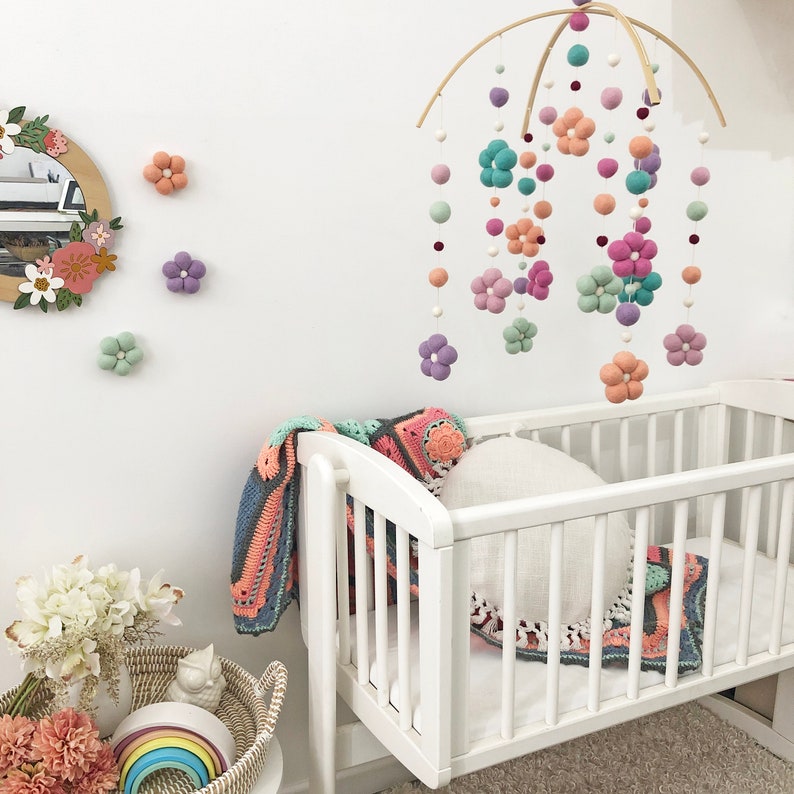 May include: A nursery mobile featuring felted wool balls and flower shapes in shades of peach, lavender, mint green, and teal, suspended from a curved wooden frame. The mobile hangs above a white wooden crib with a crocheted blanket and round pillow.