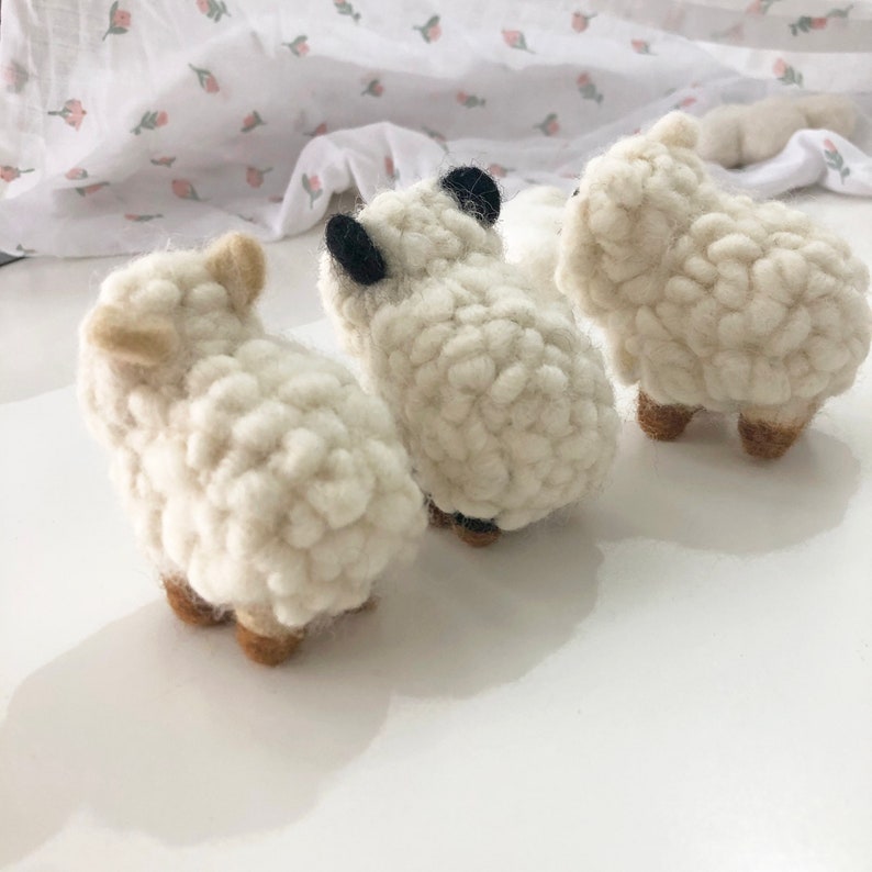 May include: Three white fluffy sheep made of felt, standing in a row. The sheep are facing the same direction, with their backs to the viewer. The sheep have black eyes and brown legs.
