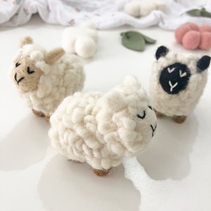 May include: Two white felt sheep toys. One sheep has black eyes and a black nose. The other sheep has brown eyes and a brown nose.
