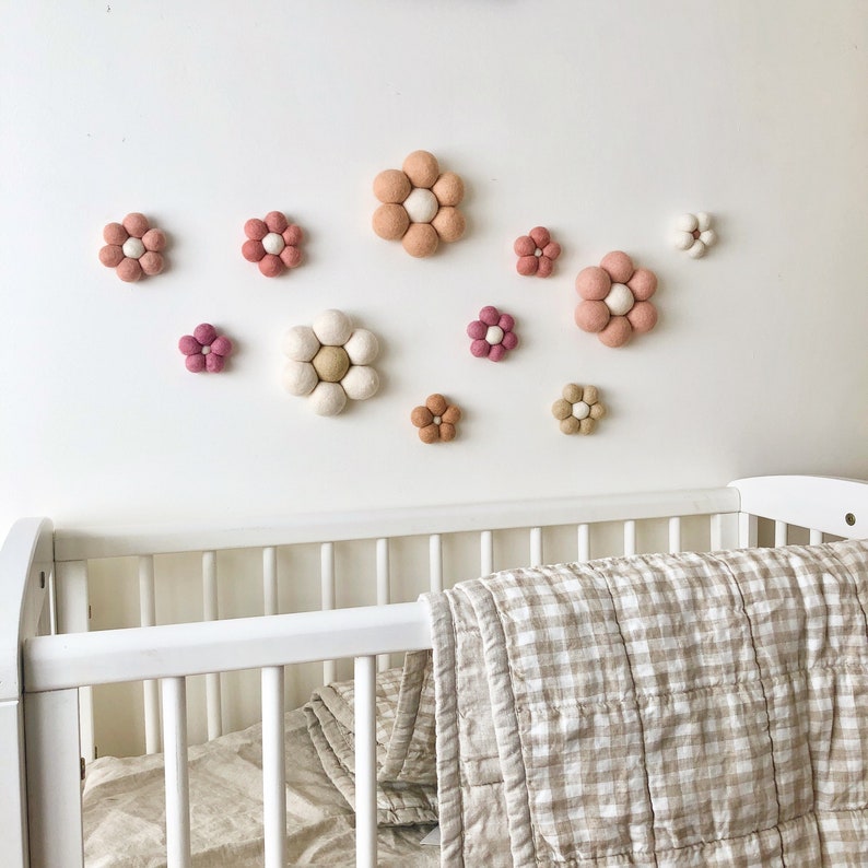 May include: A collection of felt flower wall decor in various sizes and shades of pink, white, and beige. The flowers are mounted on a white wall above a white crib with a tan and white checkered blanket.