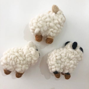 May include: Three white felt sheep toys with brown legs and black eyes. The sheep are all different sizes and are arranged in a triangular shape.