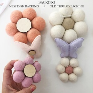 May include: A collection of felt flower and butterfly ornaments in various colors, including pink, lavender, and white. The ornaments have wooden disc backings and are displayed against a white background. The text "BACKING" and "NEW DISK BACKING / OLD THREAD BACKING" is visible.