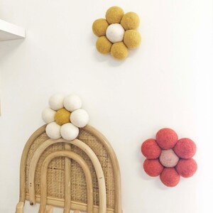 May include: Three felt flower wall decorations. One flower is yellow and white, another is yellow, and the third is red and pink. The flowers are made of round felt balls and are mounted on a white wall.