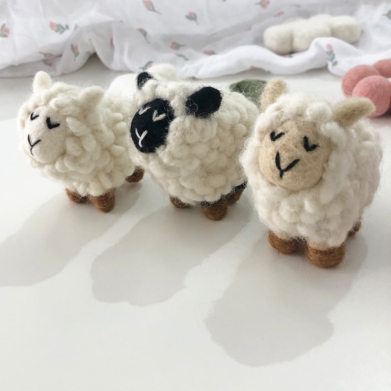 May include: Three white felt sheep toys with black eyes and brown legs. The sheep are arranged in a row on a white surface.