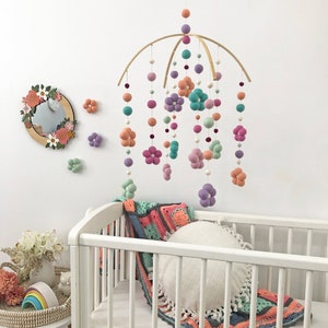 May include: A hanging mobile featuring felted wool balls and flower shapes in shades of peach, lavender, teal, and sage green suspended from a curved wooden frame, accompanied by a round mirror with floral accents and a white crib with a crochet blanket.