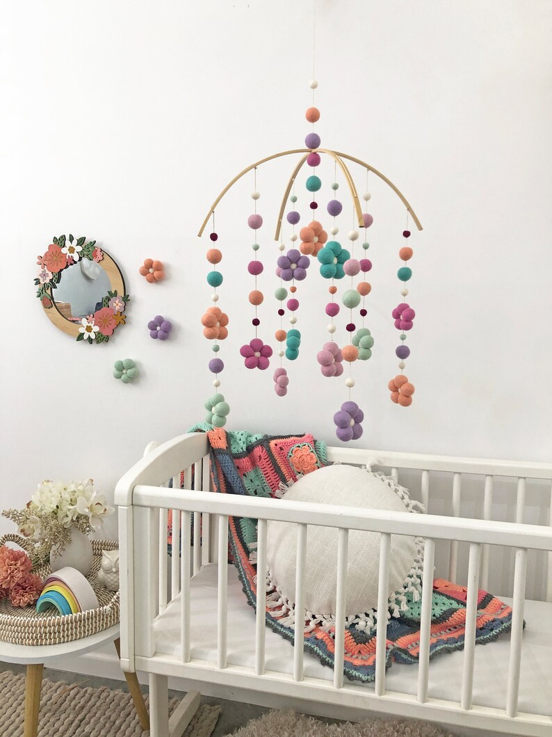 May include: A hanging mobile featuring felted wool balls and flower shapes in shades of peach, lavender, teal, and sage green suspended from a curved wooden frame, accompanied by a round mirror with floral accents and a white crib with a crochet blanket.