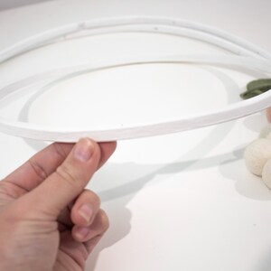 May include: A white circular wooden hoop frame for a mobile, featuring a smooth painted finish. The structure consists of two concentric rings connected by small pegs, with a small cluster of soft felt balls in muted green and cream tones attached.