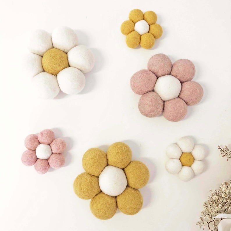 May include: A collection of felt flower decorations in various sizes and colors. The flowers feature a central sphere surrounded by petals made of smaller spheres. Colors include white, yellow, and pink, arranged against a white background.